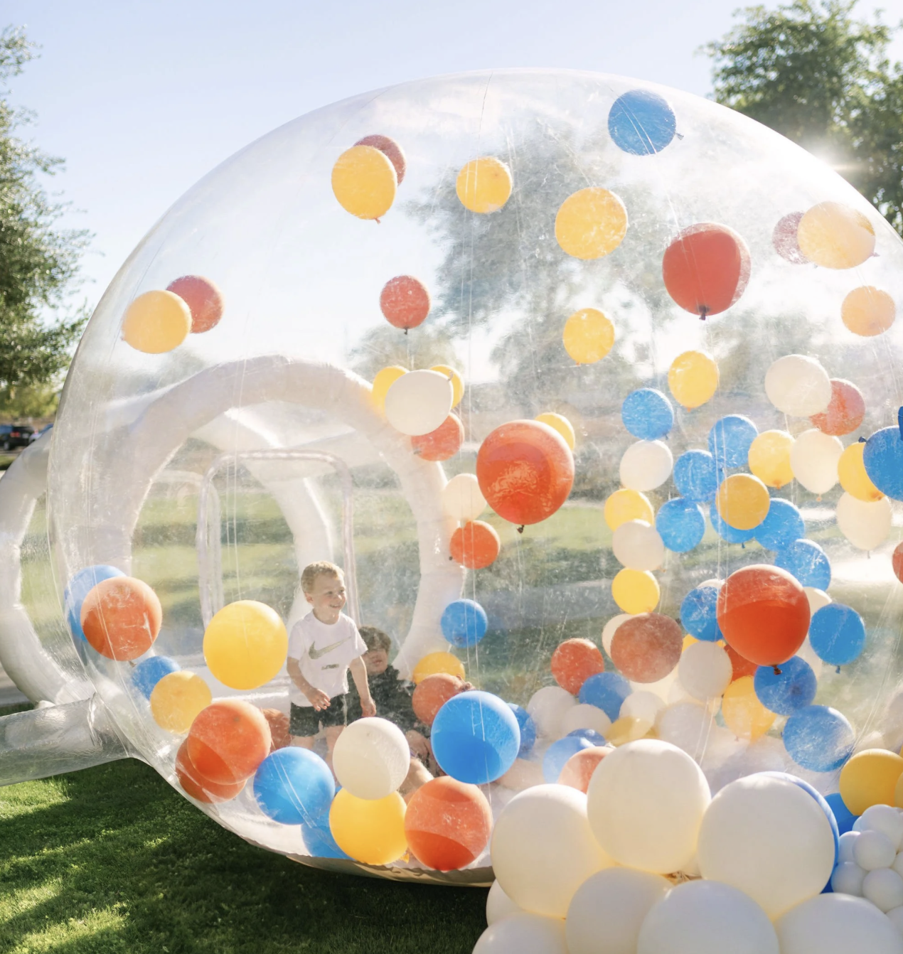 Children playing inside a large transparent inflatable sphere filled with colorful balloons on a grassy area outdoors.