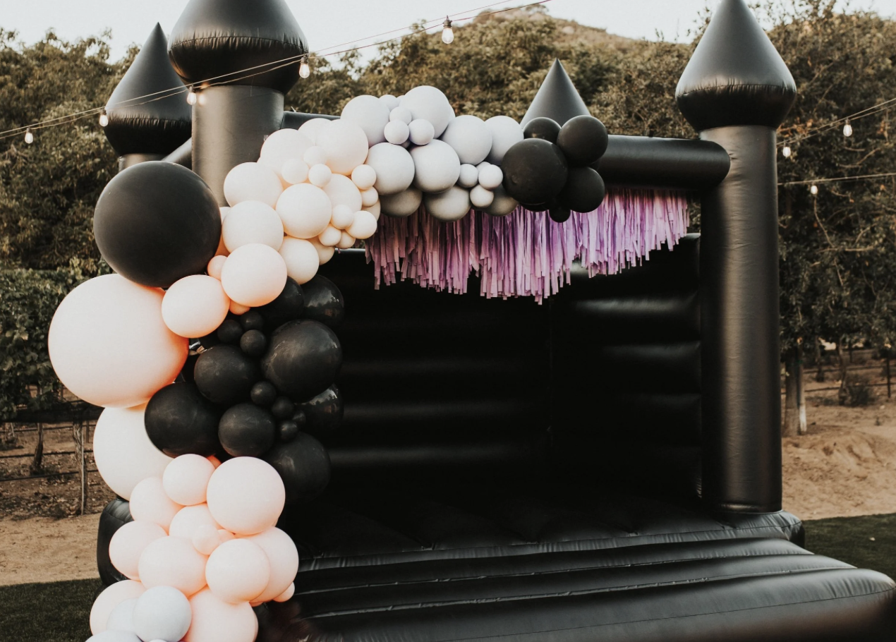 Black inflatable castle with pink fringe decoration and balloon arch in black, white, and light pink, outdoor setting with trees in background.