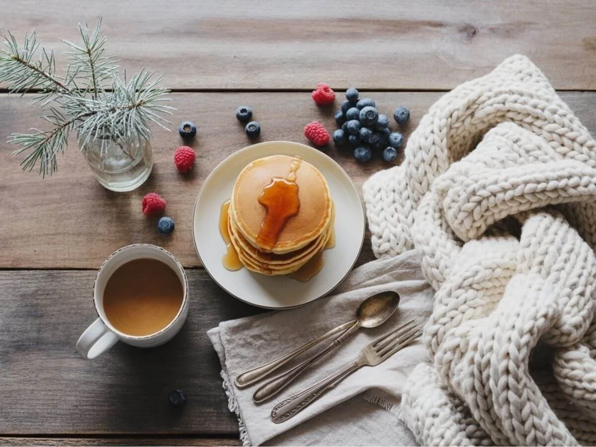 A styled flat lay photograph of an elegant winter brunch spread arranged on a rustic wooden table with soft natural lighting. T