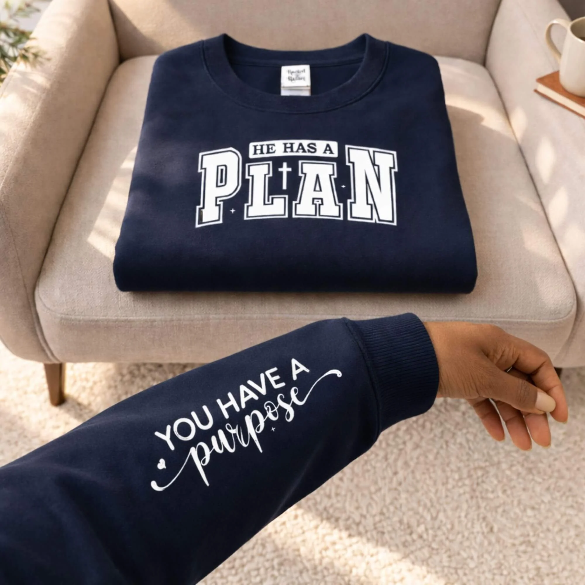 Navy blue Christian sweatshirt with the message “He Has a Plan” and a cross design, with sleeve text reading “You Have a Purpose,” displayed on a chair with a coffee mug nearby.