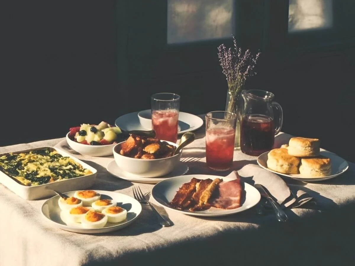 Southern-style brunch table with deviled eggs, biscuits, bacon, breakfast casserole, fried chicken, fresh fruit, and iced drinks in morning sunlight.