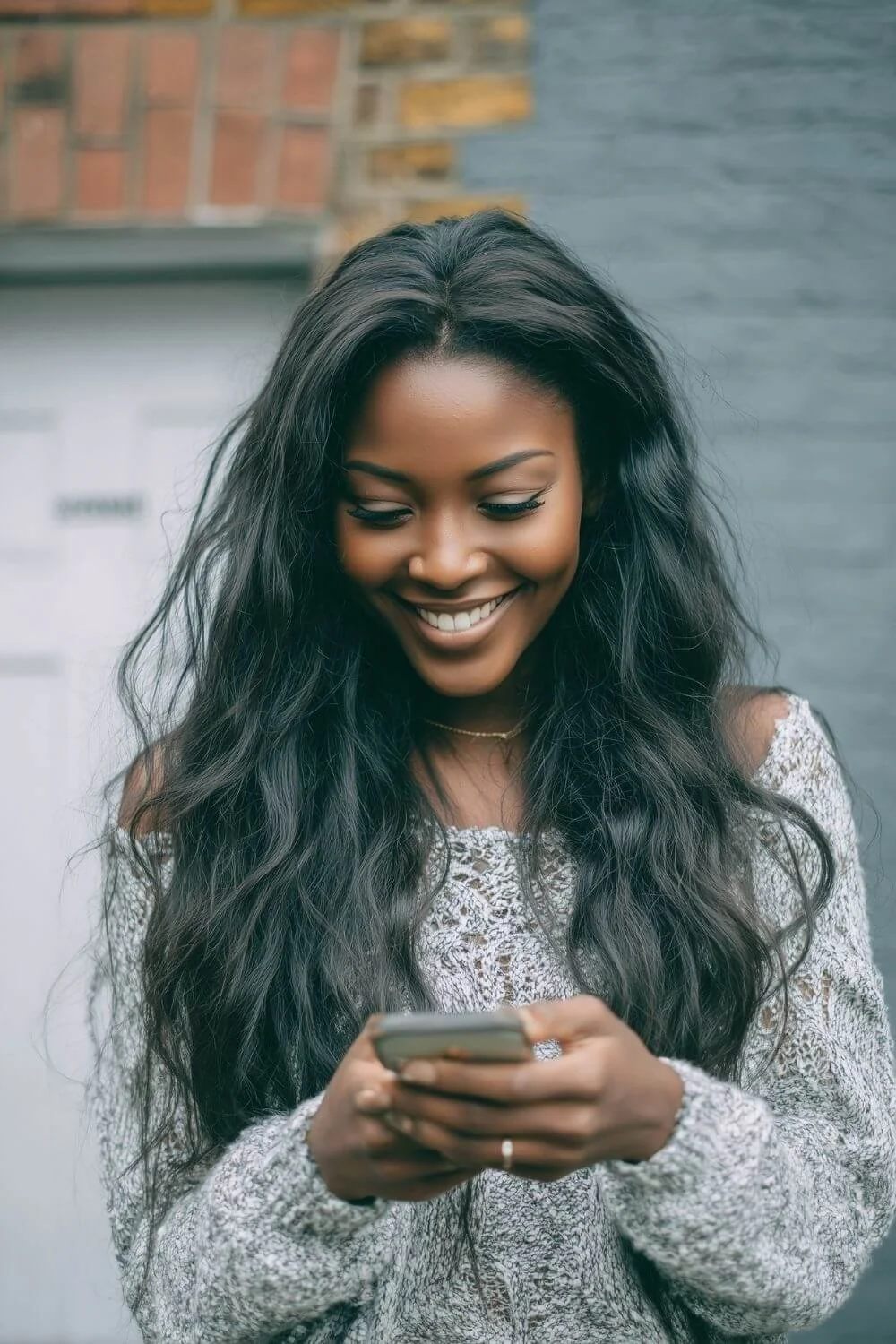 Smiling woman outdoors looking down at her phone with long wavy hair and a soft knit sweater.