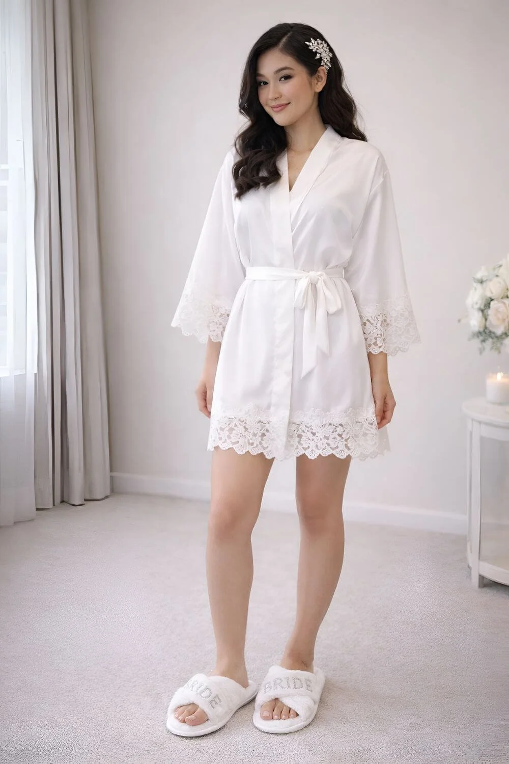 Asian bride standing in a white lace-trim satin robe and fluffy “Bride” slippers in a bright getting-ready room.