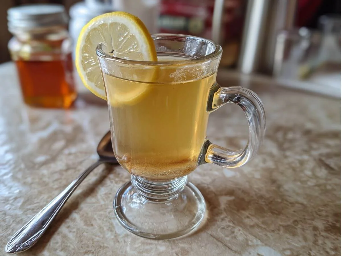 Clear glass mug of warm lemon and honey tonic on a kitchen table with a lemon slice inside.