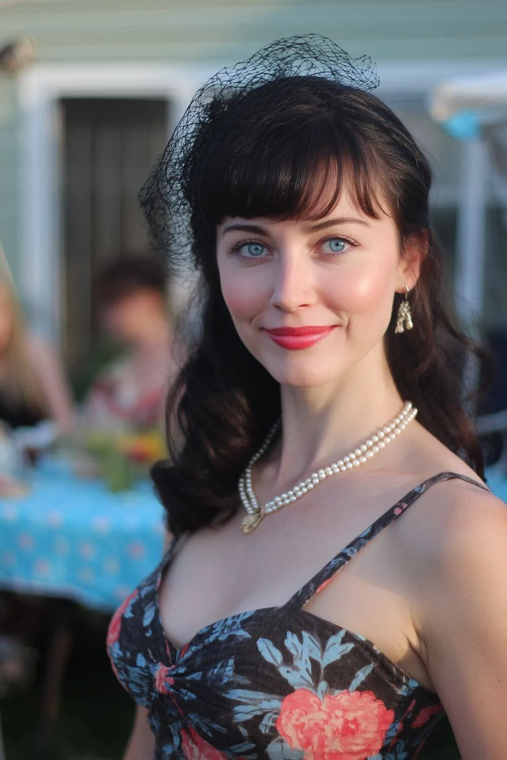 Woman with dark hair wearing a floral dress, pearl necklace, and small black fascinator hat, smiling at an outdoor garden gathering.