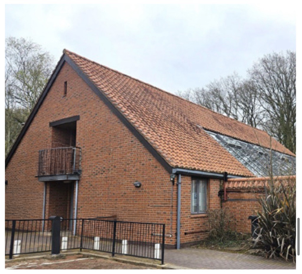 A two-story brick house with a steep gabled roof that has orange tiles. The house features a small balcony and a window on the upper level, and a window on the lower level. There is a black fence in front and some trees in the background.