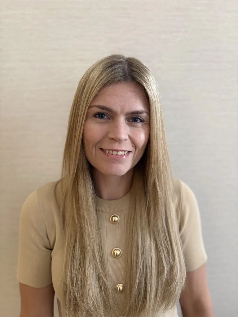 A woman with shoulder-length brown hair smiling, wearing a cream top, a navy cardigan, a pearl necklace, and a gold pin with the initials 'DL' on her cardigan, standing against a plain beige wall.