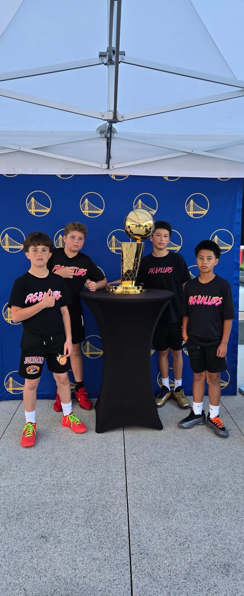 Four young boys in black basketball uniforms standing behind a black table with a large golden trophy in front of a blue backdrop with Golden State Warriors logos under a white canopy.