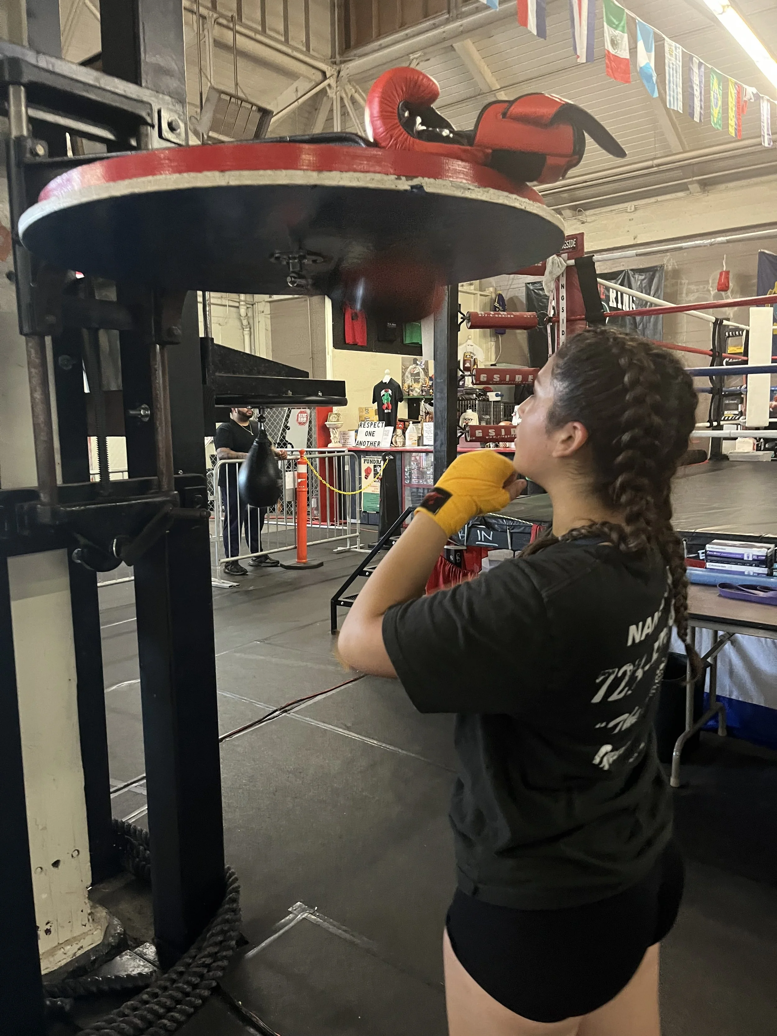 A woman in a black T-shirt and yellow boxing gloves standing in front of a red punching bag in a gym.
