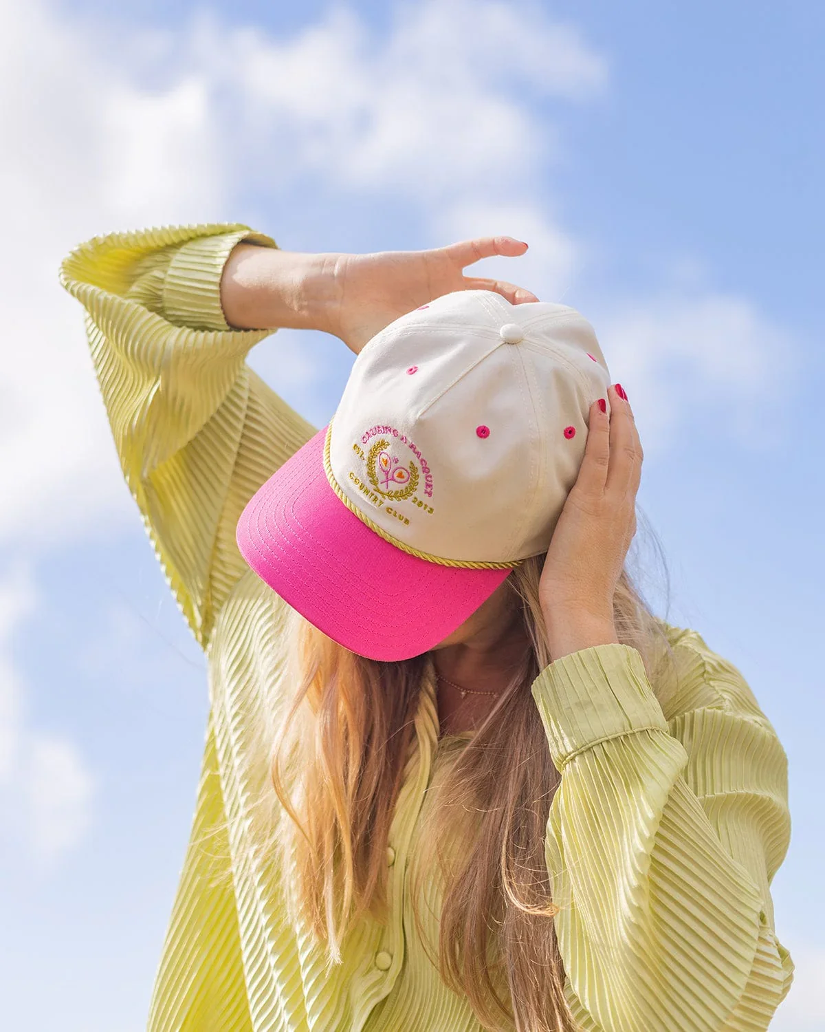 A woman with long blonde hair wearing a yellow striped shirt and a pink and white cap, covering her face with her hands, outdoors against a blue sky with some clouds.