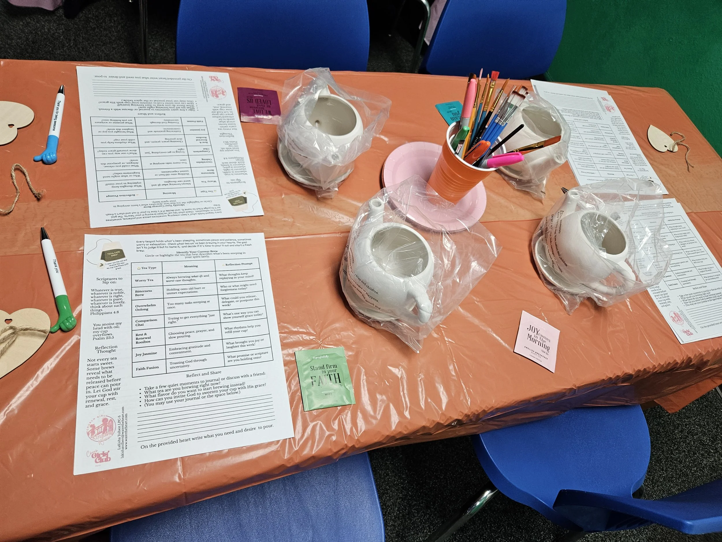 Table covered with an orange plastic tablecloth, holding papers, pens, tea bags, and three ceramic teapots wrapped in plastic. There are two white cups, also wrapped in plastic, and a container with colored pens. Blue chairs are visible around the table, with a green wall in the background.