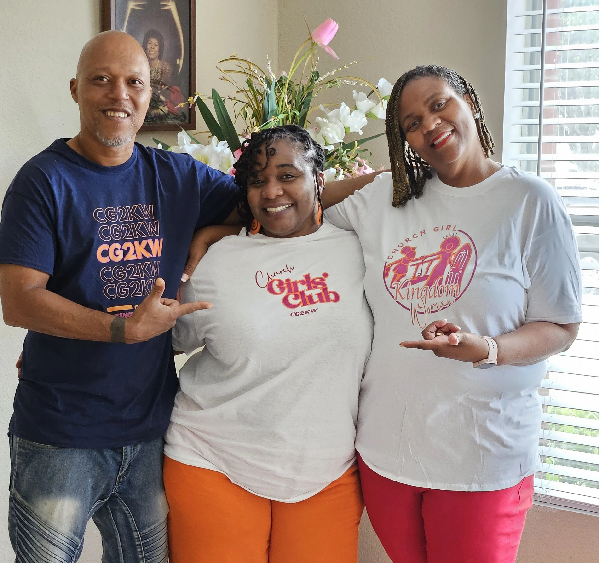 A group of three smiling adults standing indoors near a window with blinds, with a flower arrangement and a framed picture on the wall behind them. The man on the left is wearing a navy T-shirt with text, the woman in the middle is wearing a white T-shirt with 'Girls Club' printed on it, and the woman on the right is wearing a white T-shirt with pink graphics.