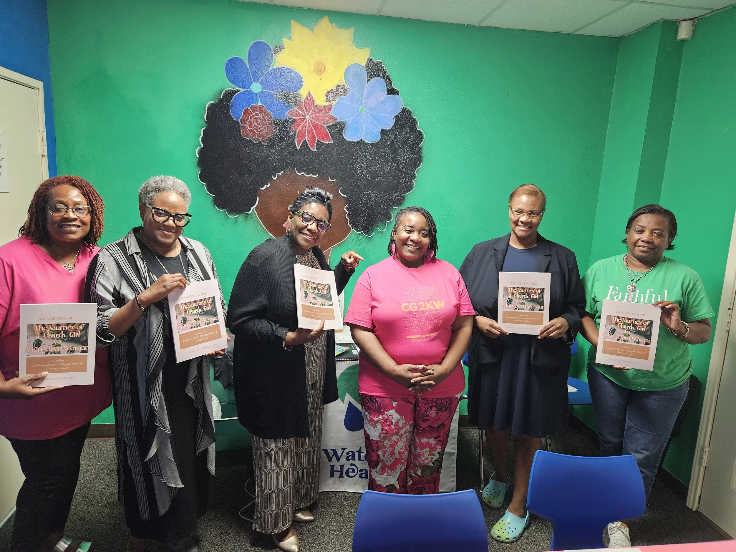 Six women standing indoors, smiling, holding flyers, in front of a colorful mural of a woman with an afro decorated with flowers.