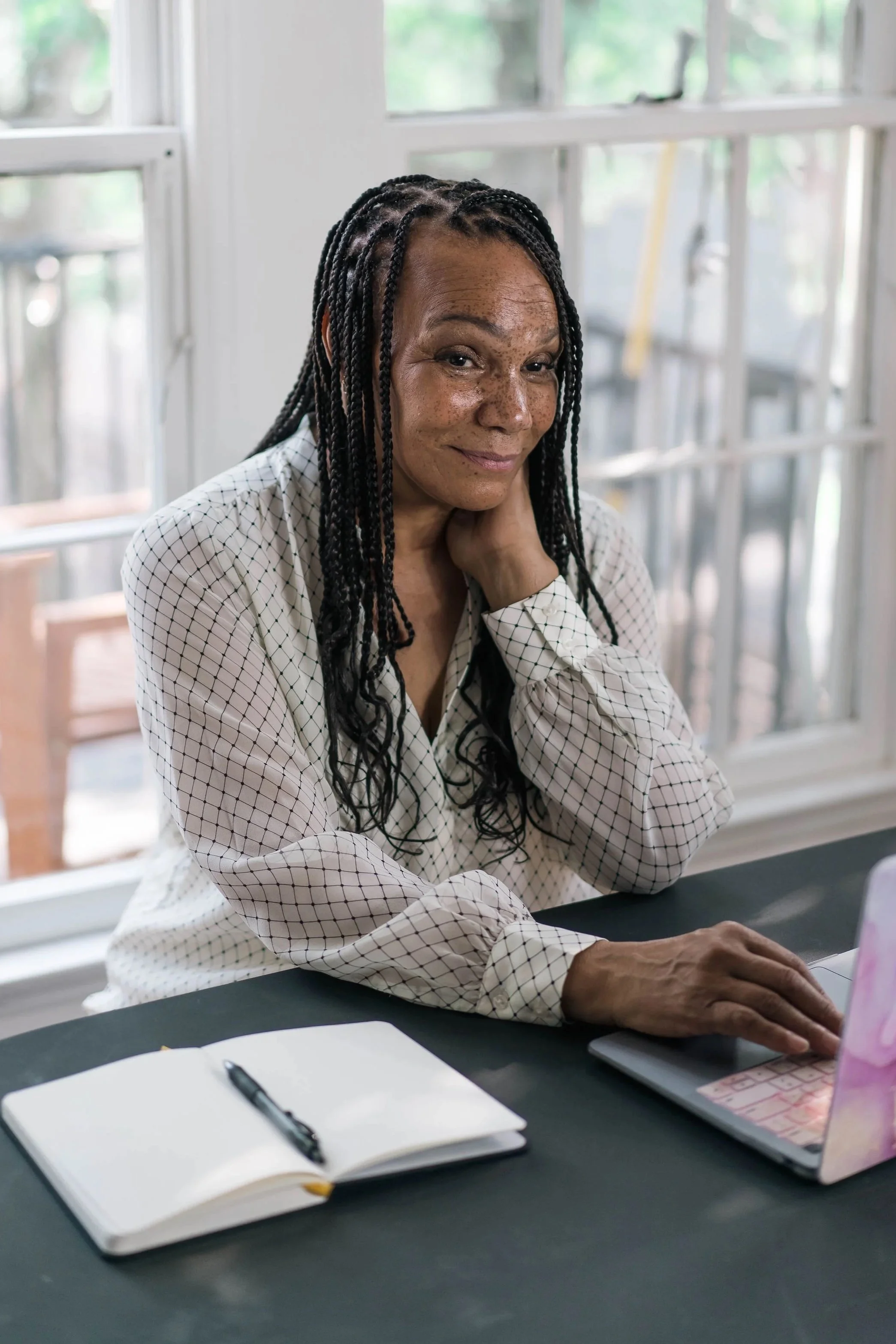 An older woman with dark braided hair and freckles is sitting at a desk in front of a laptop, smiling softly. She is resting her left hand on her neck and her right hand on the desk. The desk has an open notebook, a pen, and some papers. Behind her, there are large windows with a view of trees outside.