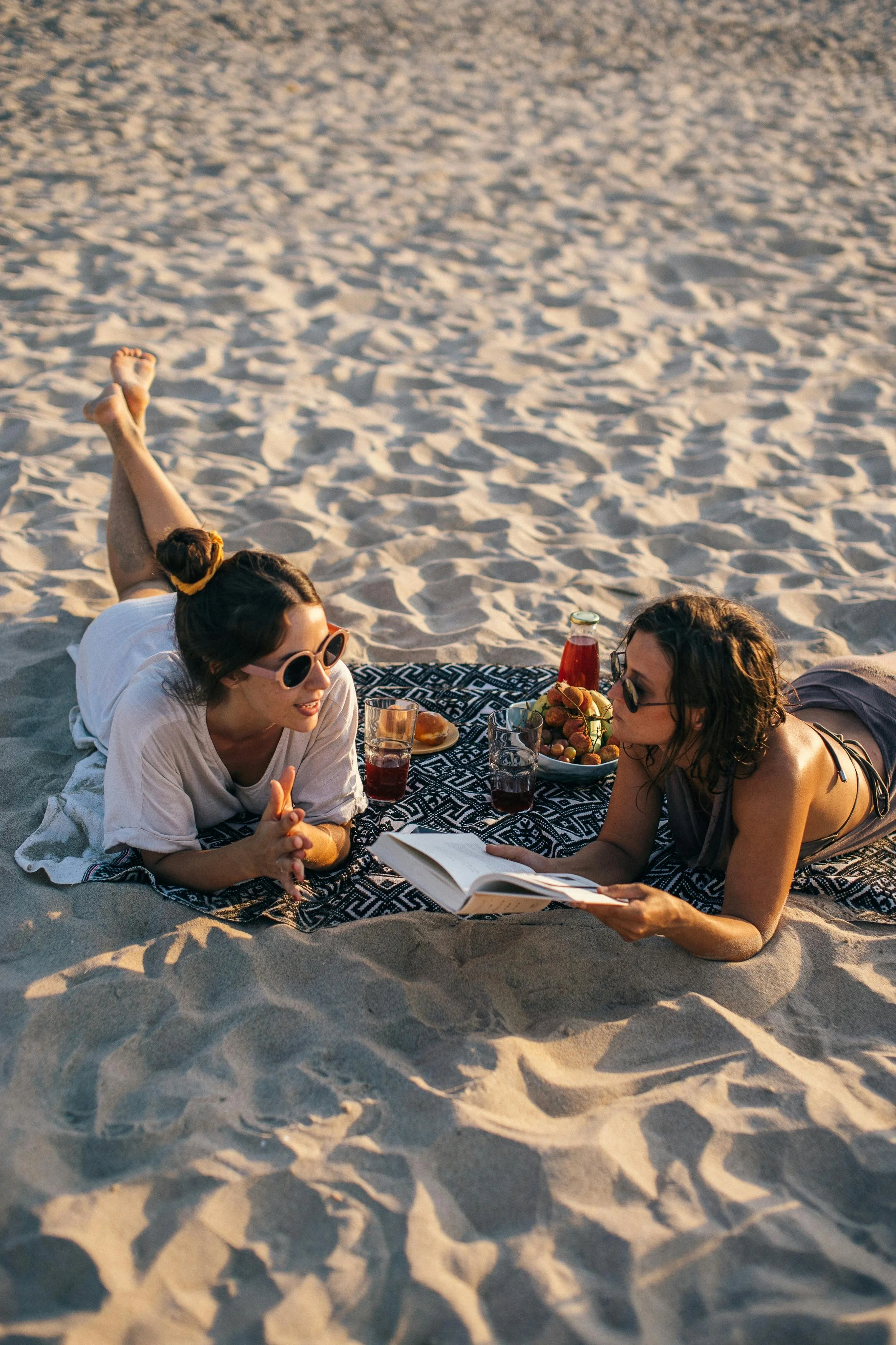 Three women are lying on the sand at the beach, engaging in a conversation. One woman is reading a book, while there are snacks and drinks on a blanket nearby.