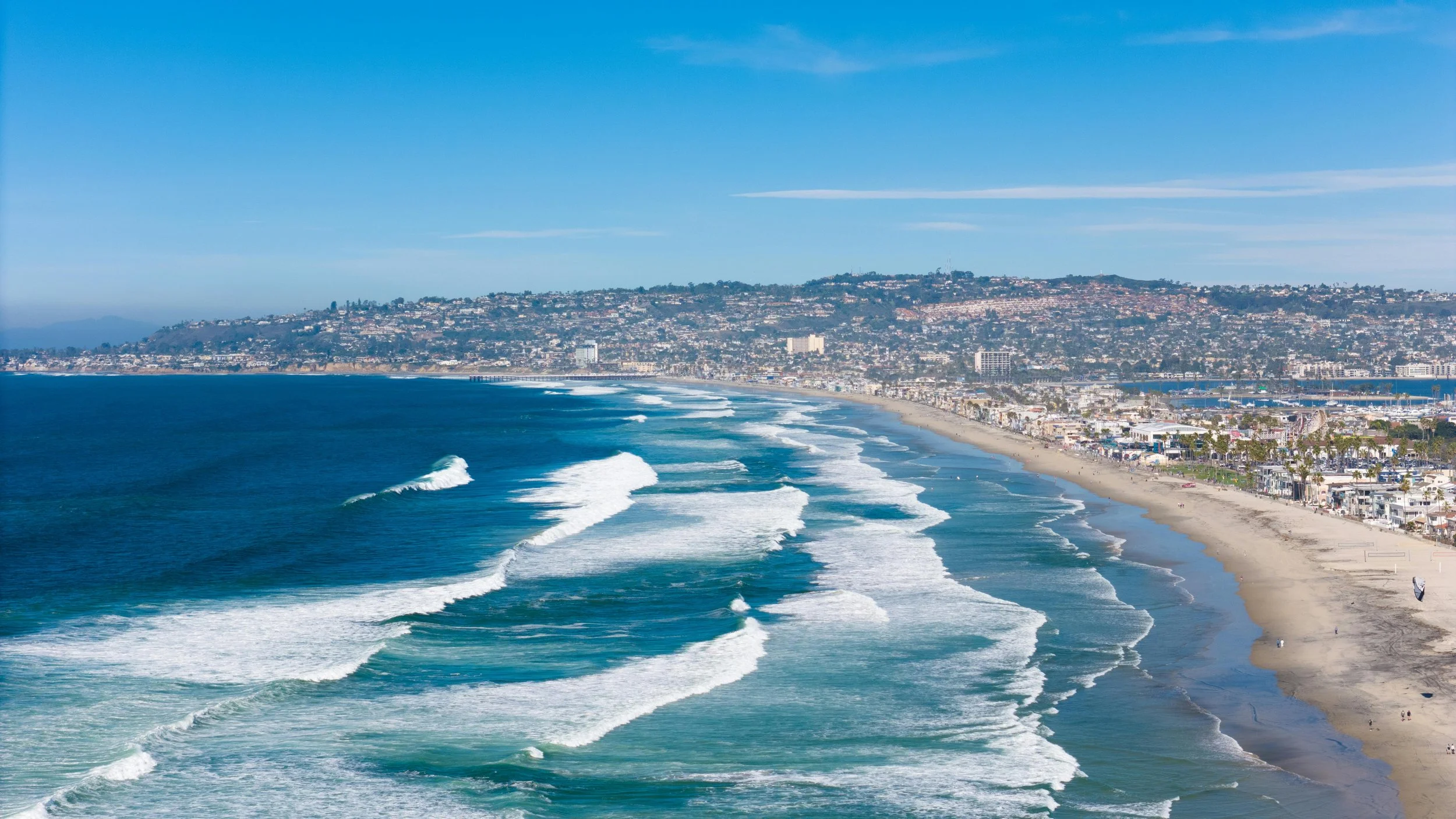 Aerial view of a beach with white waves, sandy shore, and a cityscape with buildings stretching into the hills under a blue sky.