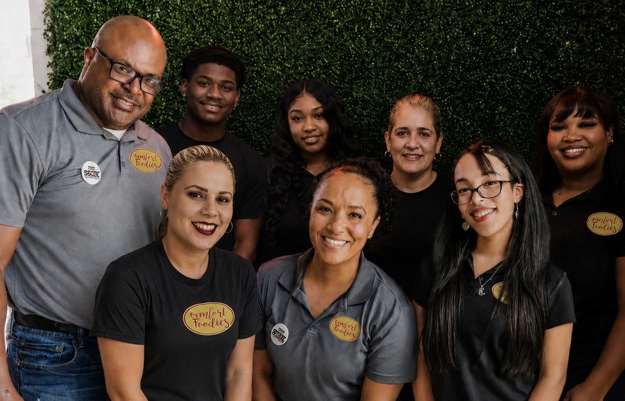 Group of nine diverse people smiling, wearing black and gray shirts with badges, standing in front of a green leafy background.