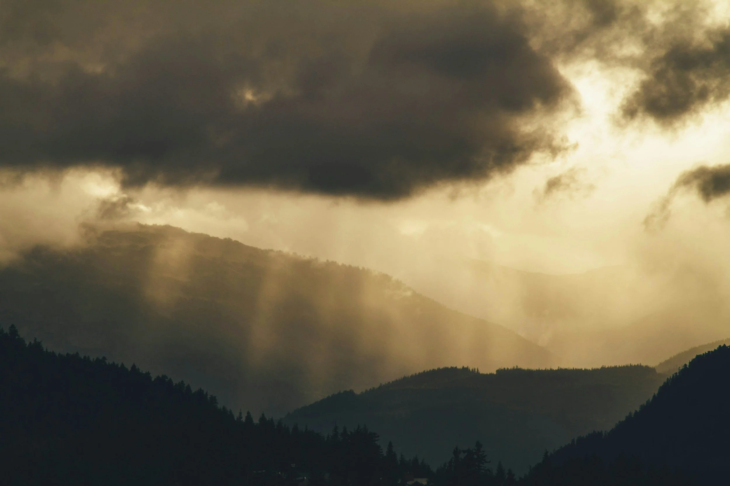 Mountain landscape with dark, stormy clouds and sun rays breaking through in a hazy, atmospheric scene.