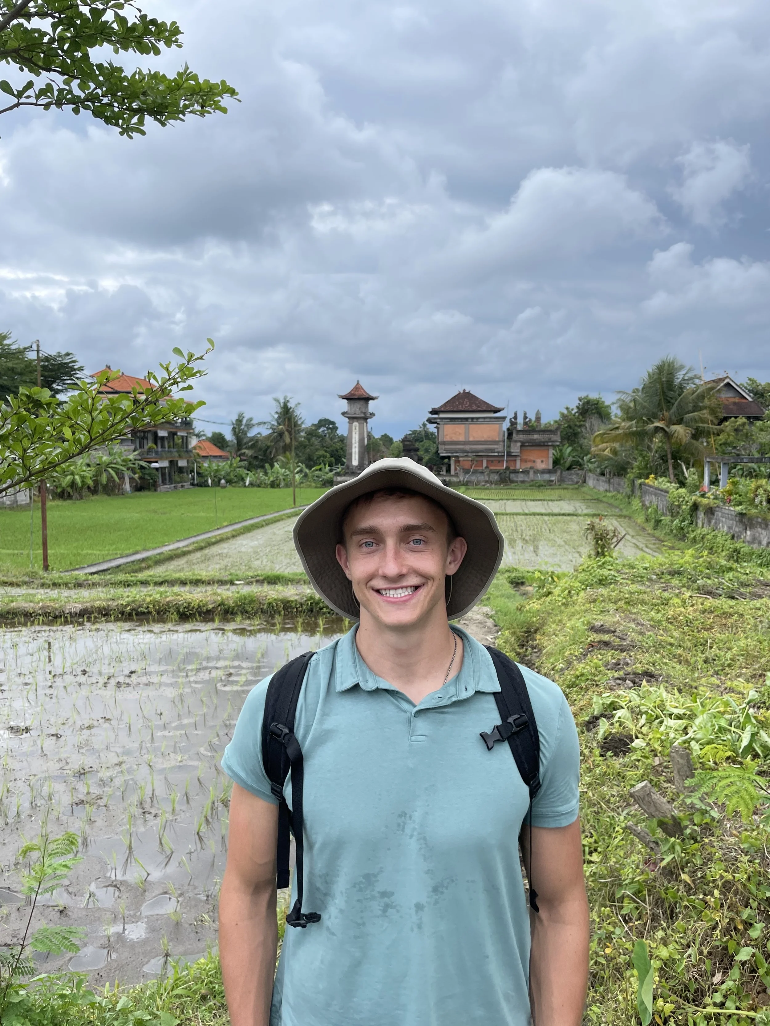 A young man smiling in a rural rice field with traditional buildings and a cloudy sky in the background.