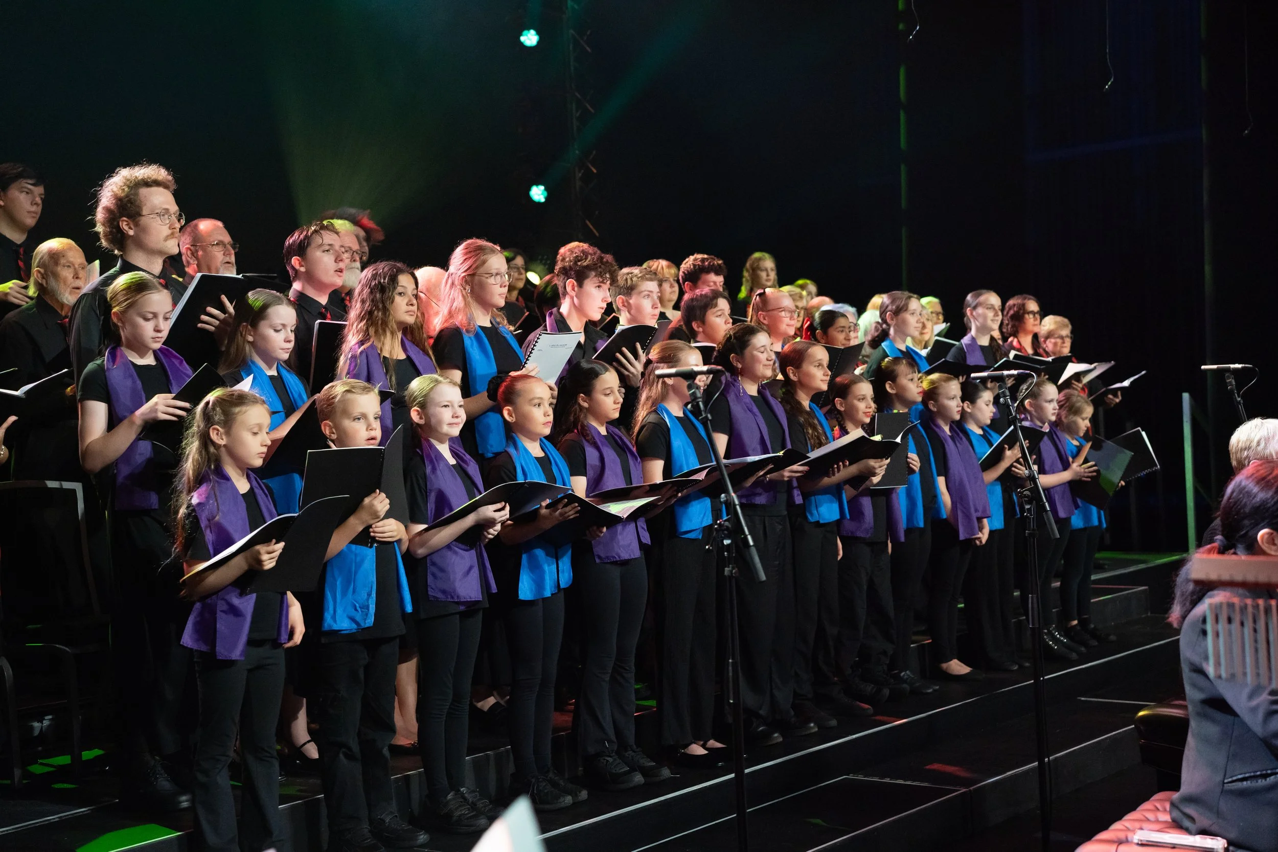 Children and adults singing in a choir on stage during a performance, many holding songbooks and wearing black clothing with purple and blue scarves, stage lighting illuminating the scene.