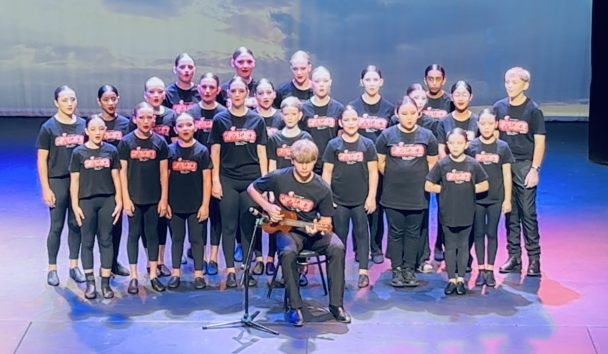 A group of children and teens wearing black T-shirts with red and white text, standing on stage with a young boy playing a guitar in front of them. The stage has a backdrop with a scenic design.