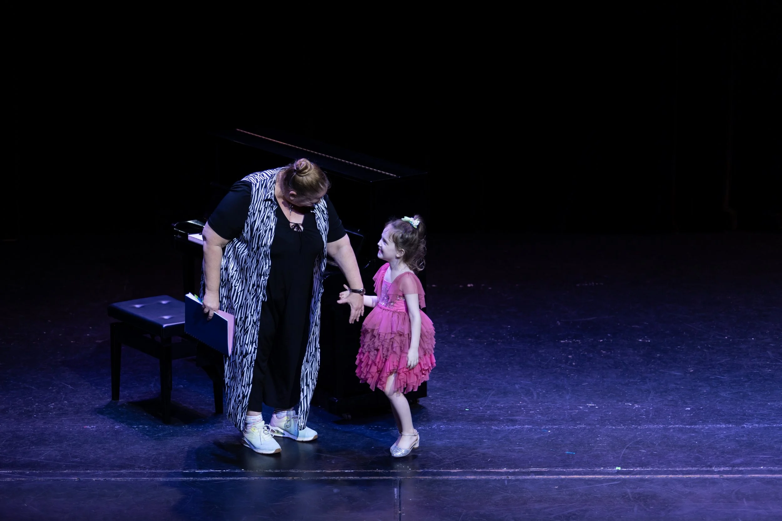 A young girl in a pink dress and a woman in black with a striped vest standing on a stage near a piano, holding hands and smiling at each other.