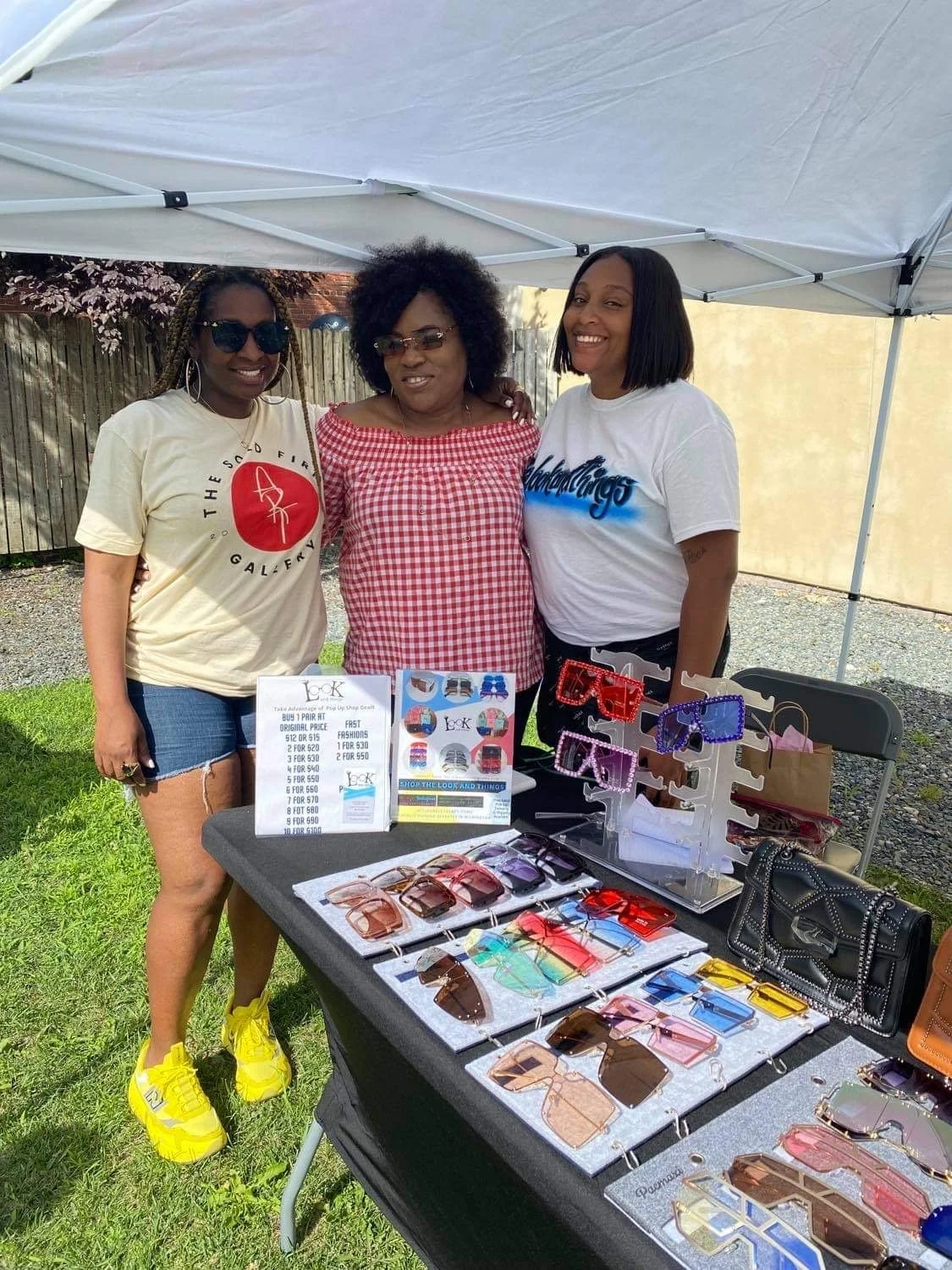 Three women standing under a white tent next to a table displaying various sunglasses and a few handbags outdoors, with a wooden fence in the background.