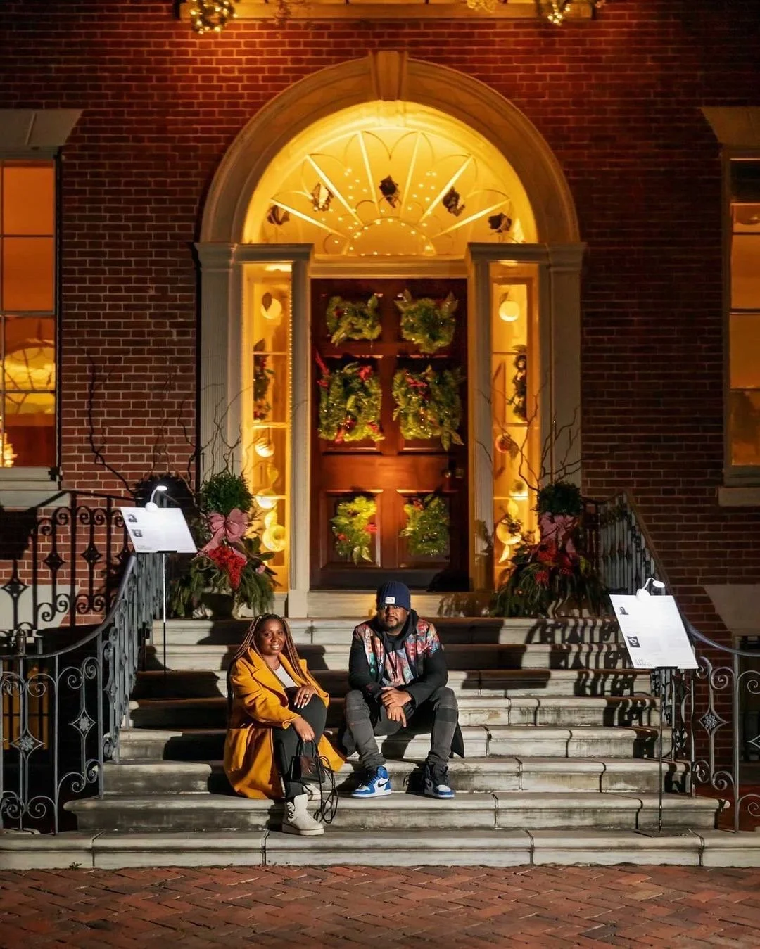 Two people sitting on the steps of a brick building entrance adorned with wreaths, under warm lighting. There are decorative iron railings and informational boards on either side of the stairs.