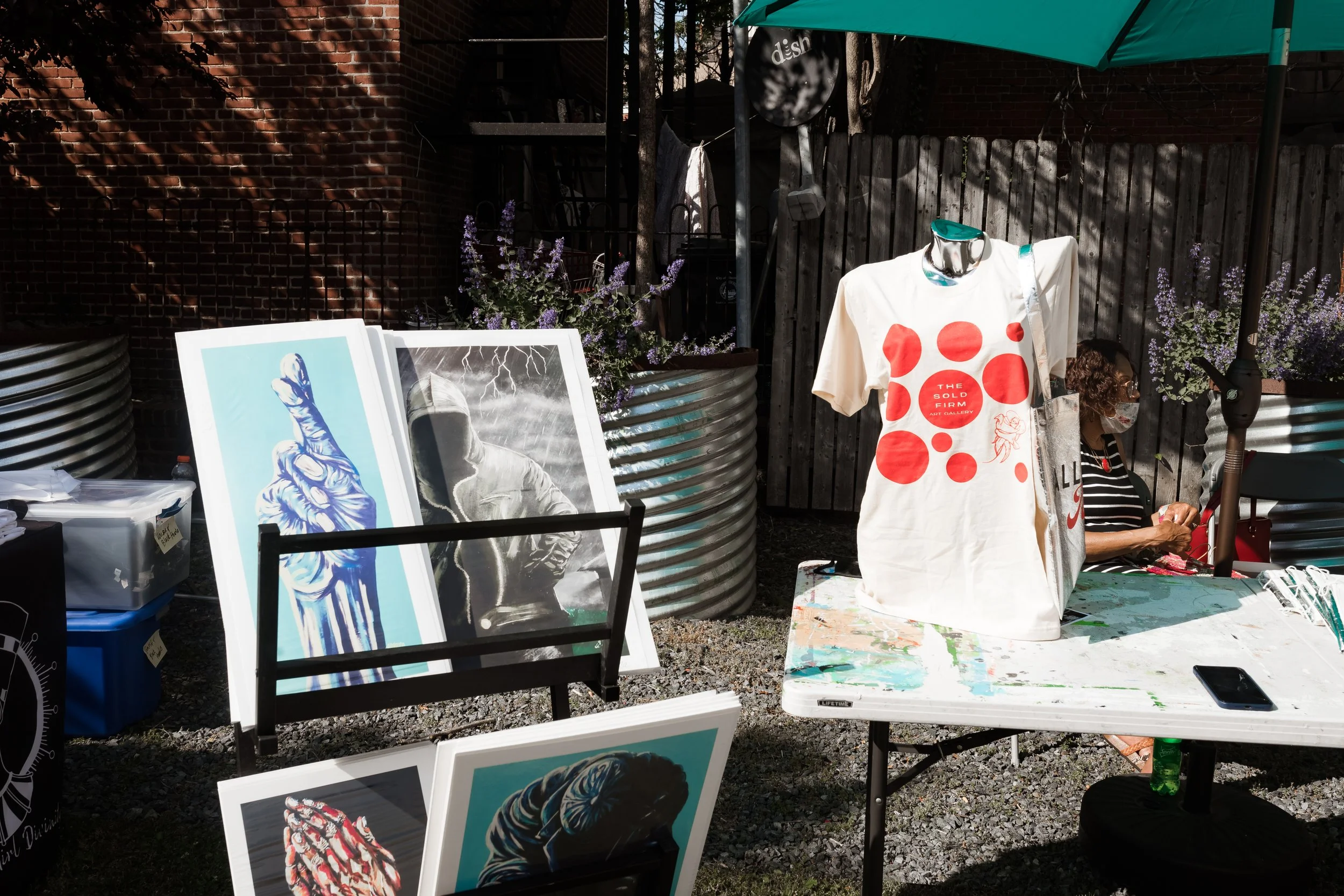 Outdoor art display featuring paintings and a white T-shirt with red circle design on a mannequin. The setting includes a table with art supplies and a seated person under an umbrella, surrounded by plants and brick fencing.