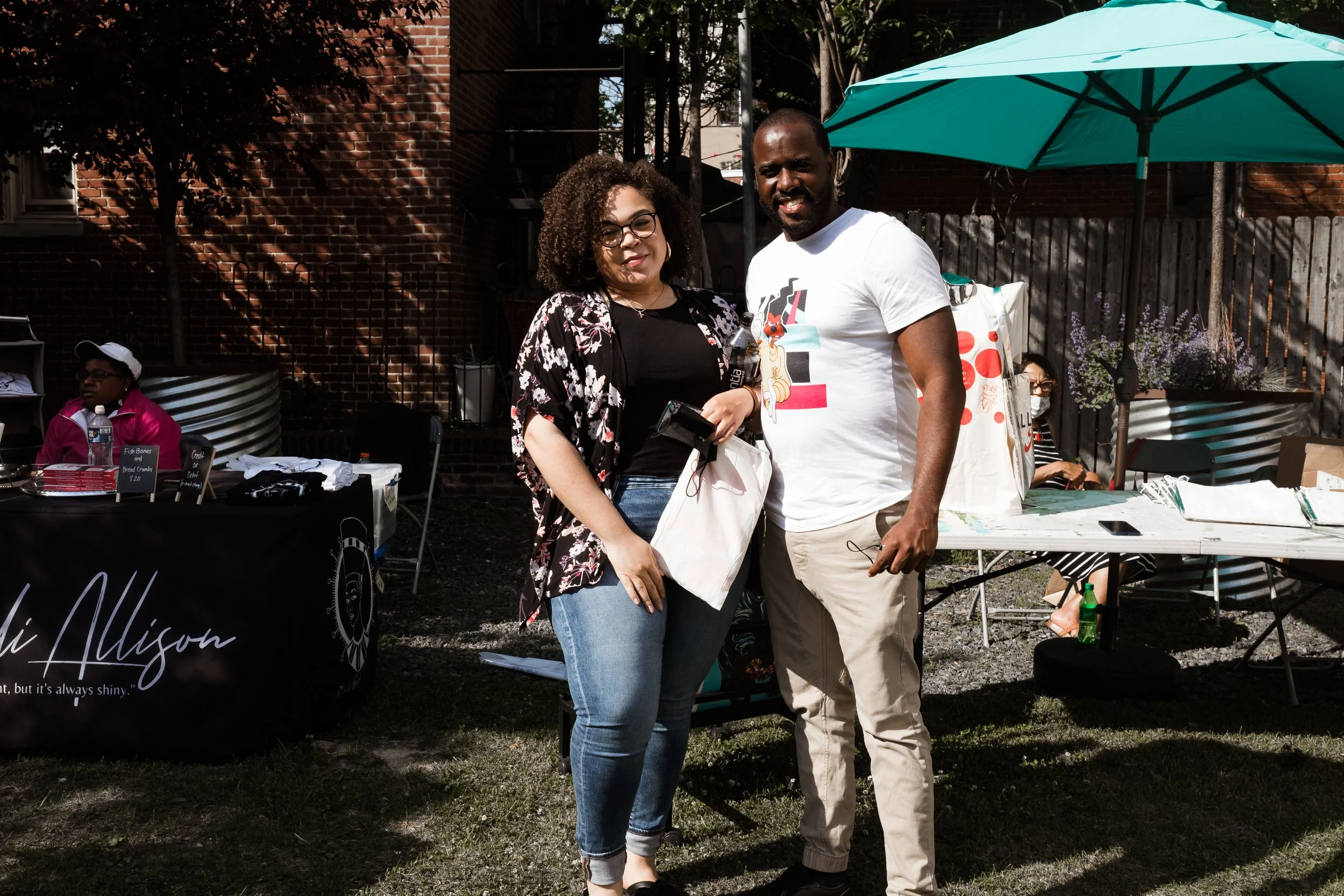 Two people posing in an outdoor setting, with small tables and a vendor display in the background. A person is seated at one of the tables, which has a teal umbrella. The scene includes a brick wall and a shady area.