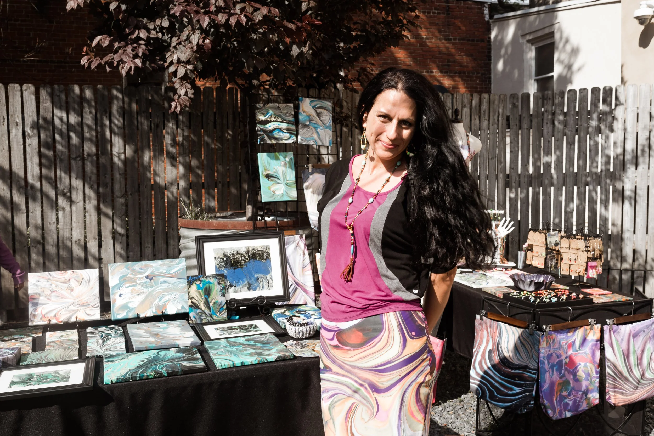 Woman at outdoor art display with abstract paintings and accessories on tables, wooden fence background.