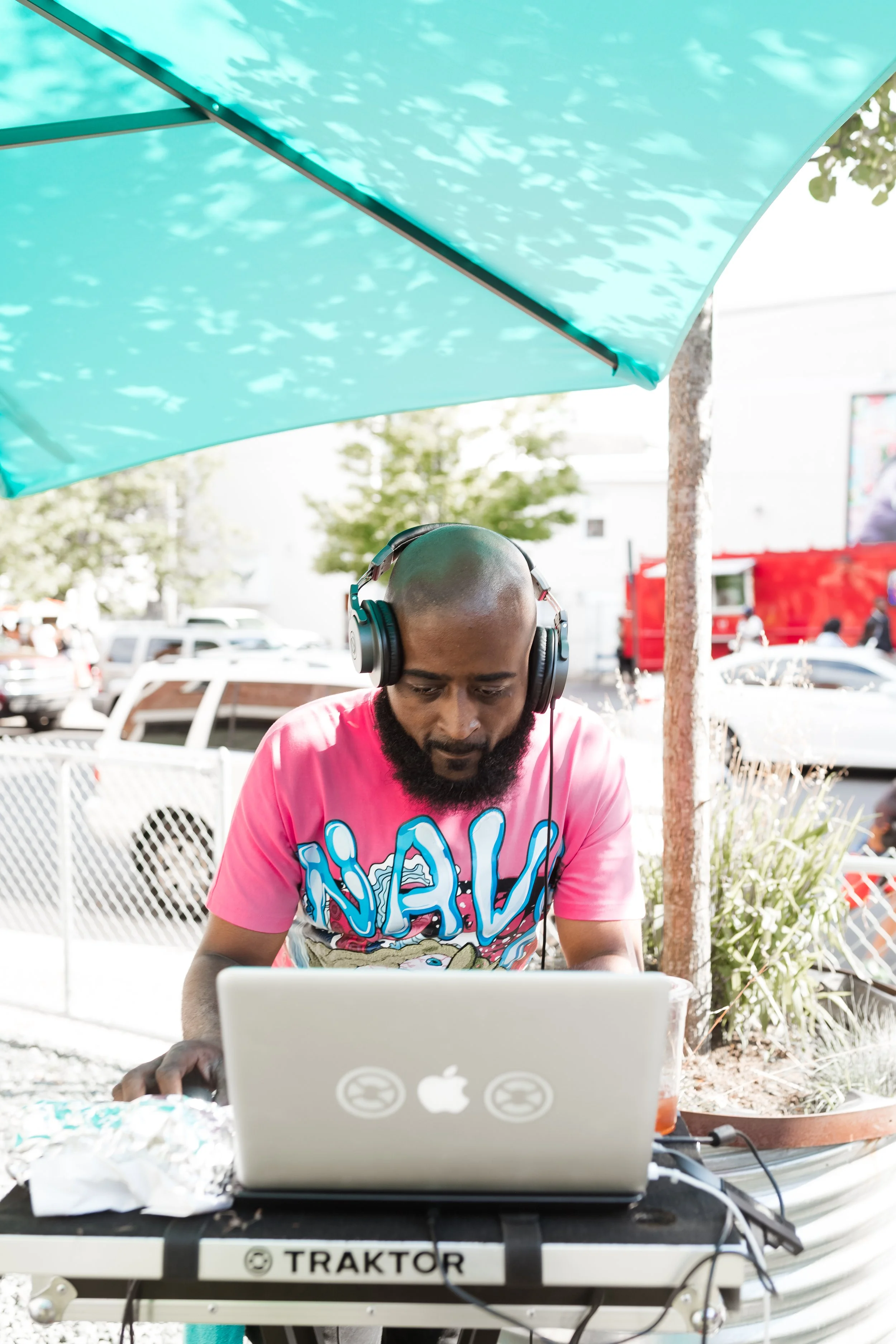 DJ wearing headphones and a pink graphic T-shirt, using a laptop outdoors under a turquoise umbrella.