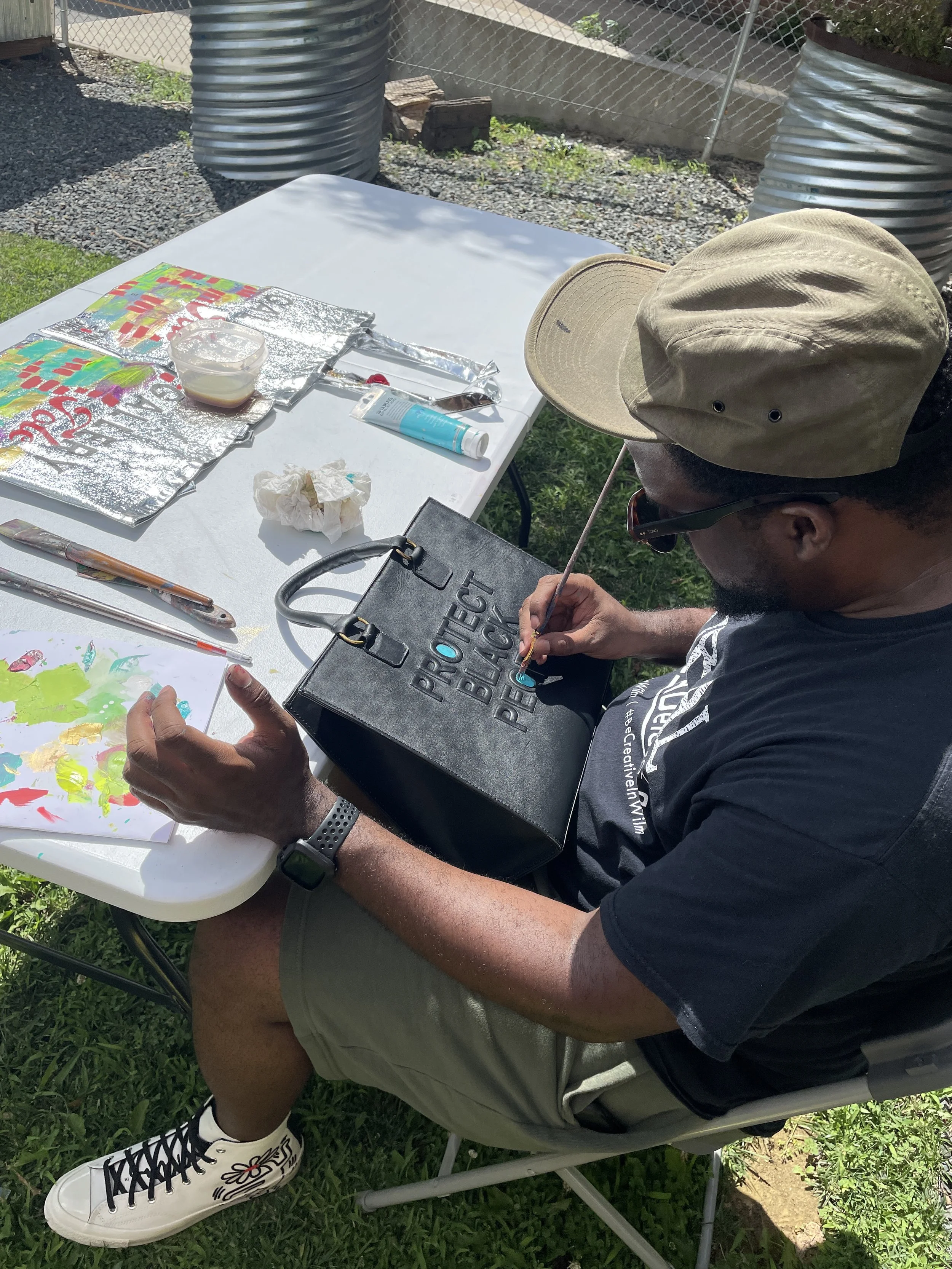 Person sitting outdoors painting a black bag at a white table with art supplies around.
