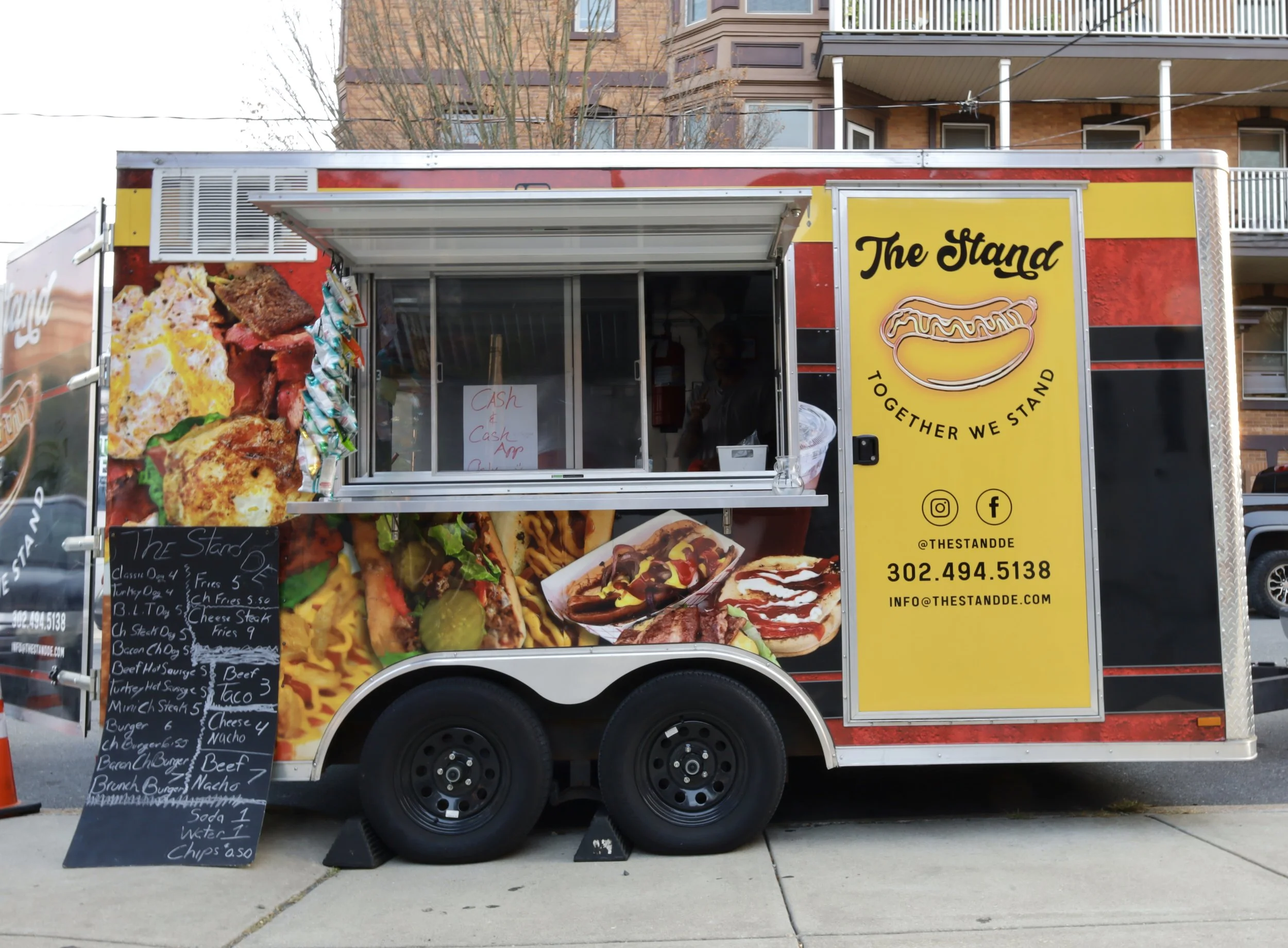 Food truck named "The Stand" with a yellow and black exterior, featuring images of food items like hot dogs and sandwiches. A chalkboard menu is displayed at the side, listing various food options. The truck offers cash and app payment options.