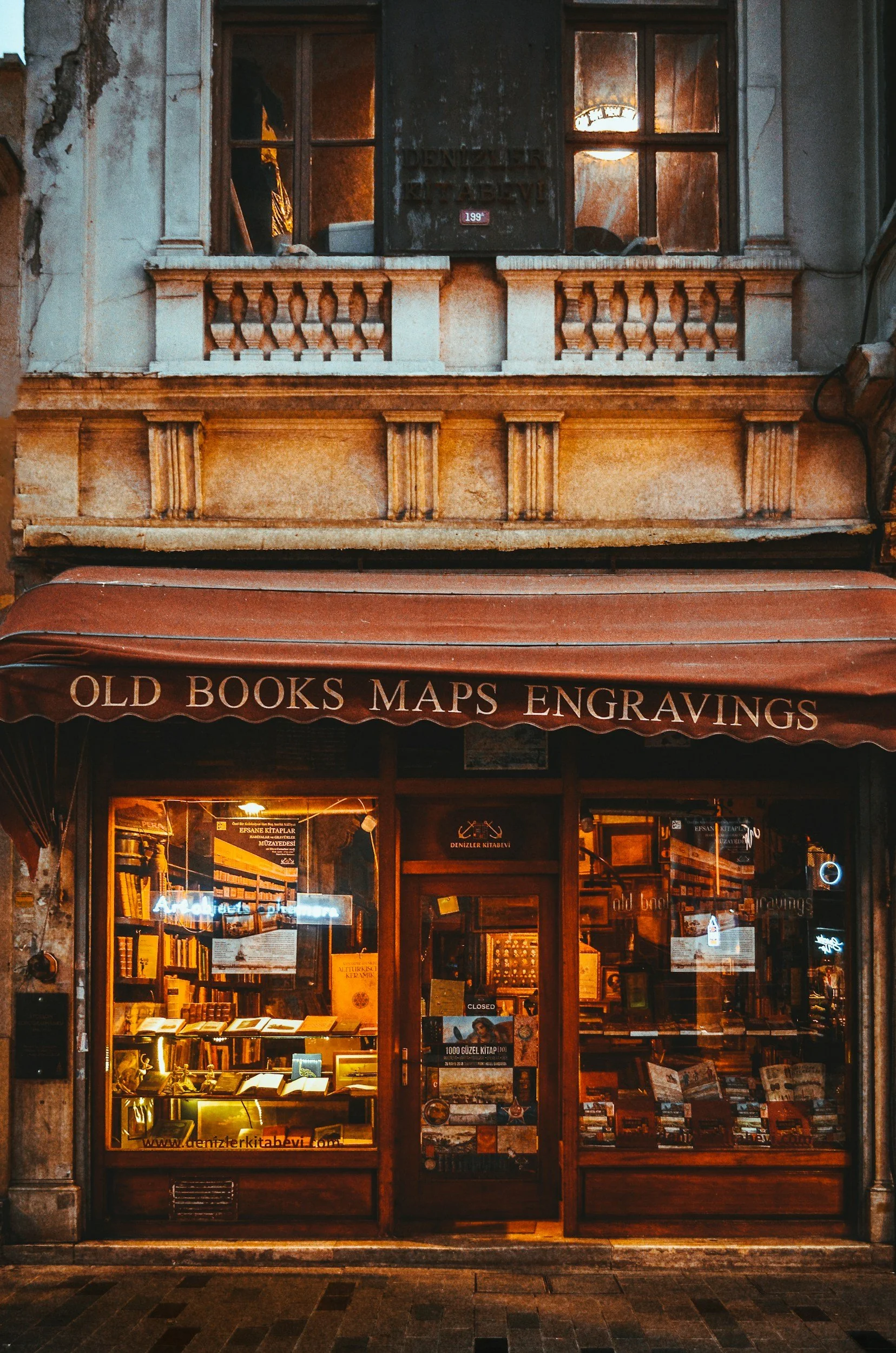 A bookstore storefront with a brown awning that reads "OLD BOOKS MAPS ENGRAVINGS." The shop has large glass windows displaying books and illuminated by warm lighting, with posters and signs on the door and window.