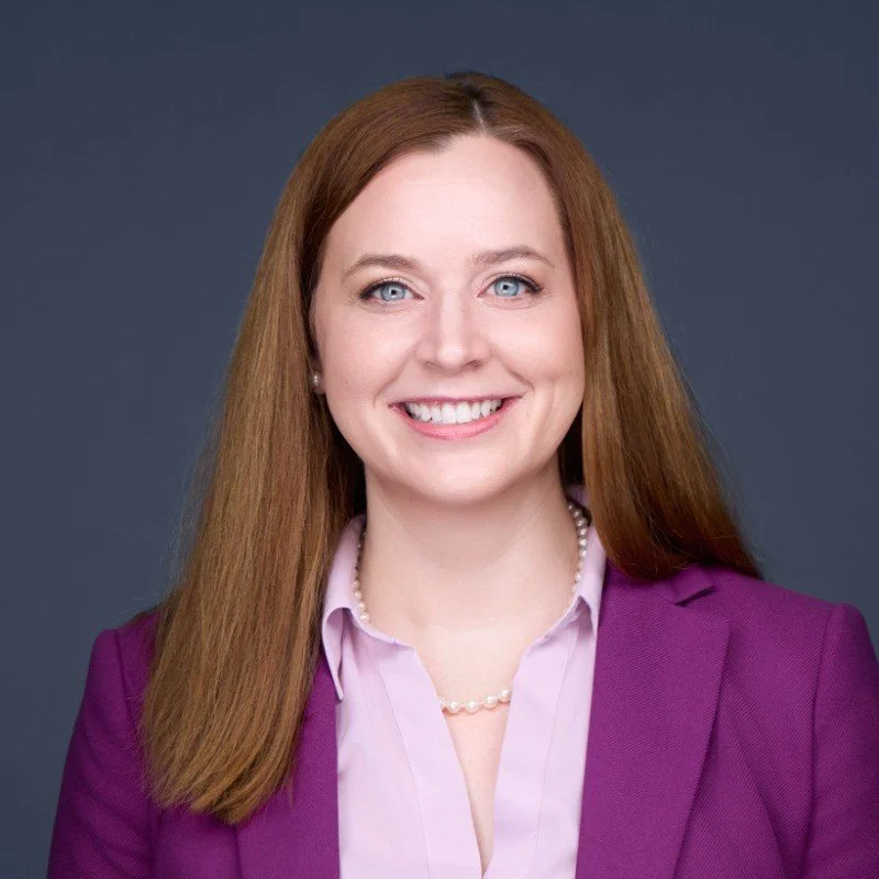 A woman with shoulder-length light brown hair wearing a white shirt, black blazer, and pearl necklace standing outdoors in a park with greenery.