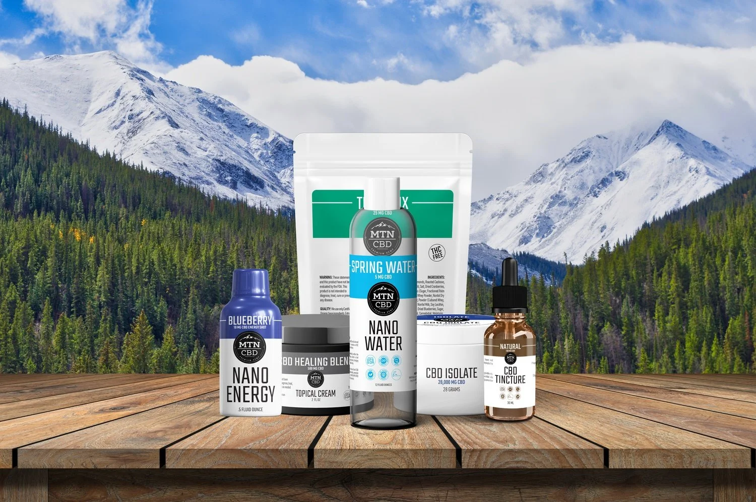 Collection of CBD products including energy shot, topical cream, spring water, nano water, isolate, and tincture on wooden surface in mountain landscape background.