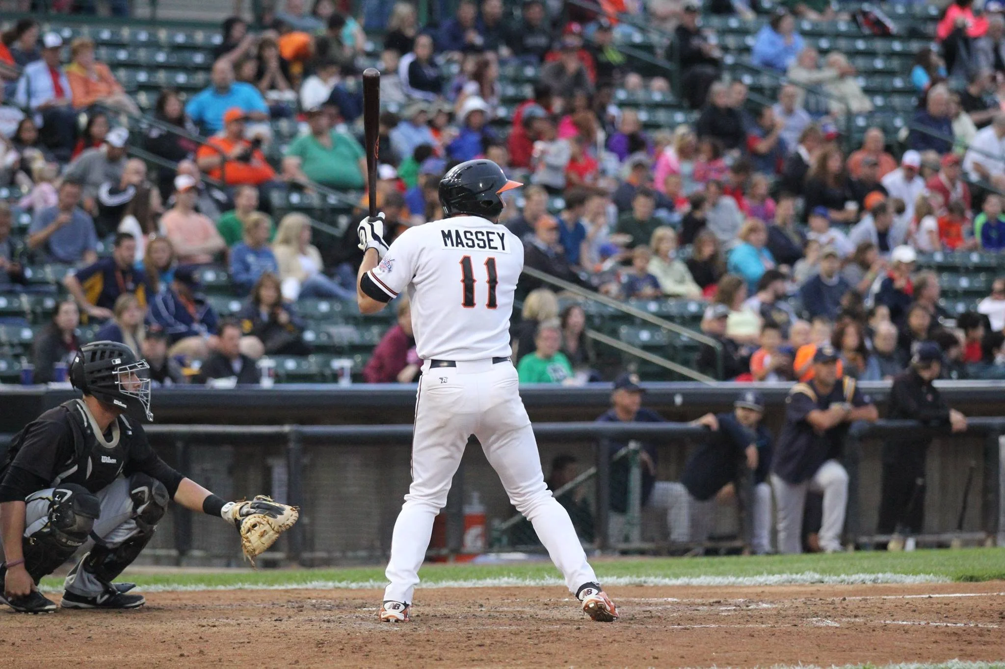 Baseball player wearing jersey with number 11 and name Massey prepares to swing at pitch, with catcher crouched behind home plate and spectators in the background