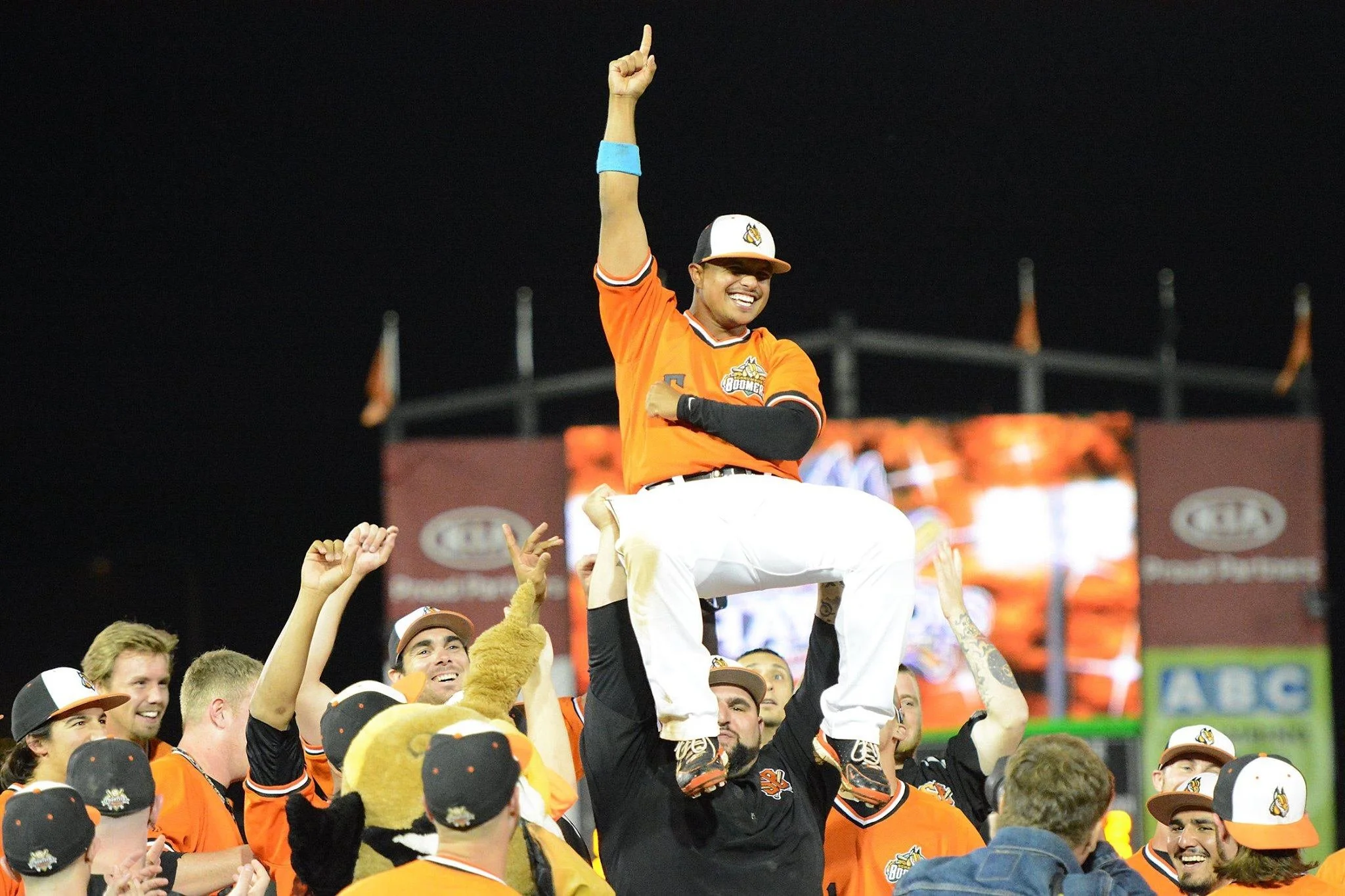 Baseball team celebrating on the field with players raising a trophy high in the air as confetti flies.