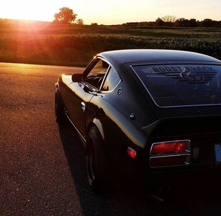 A black sports car parked on the side of a road during sunset, with a rural field in the background and a windshield decal that says 'BOOST SWAGZ'.
