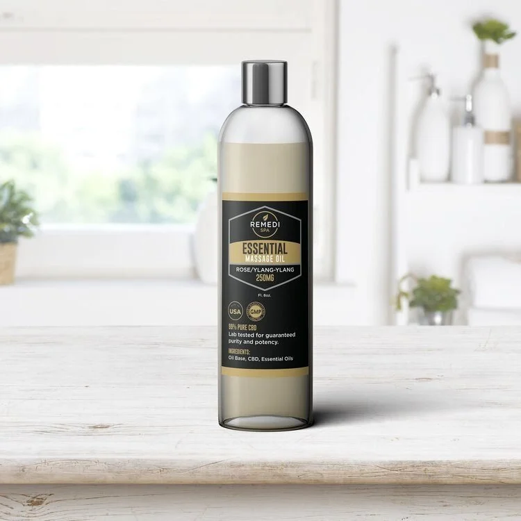 A bottle of Remedi Spa Essential Massage Oil with rose and ylang-ylang scent, containing 250mg CBD, on a white wooden surface with a blurred bright kitchen background.