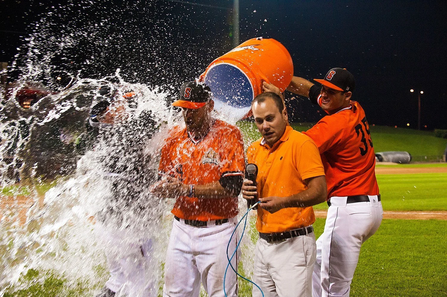 Baseball players in orange and black uniforms celebrating on the field at night, one pouring water on a teammate, with a man in a polo shirt holding a microphone nearby.