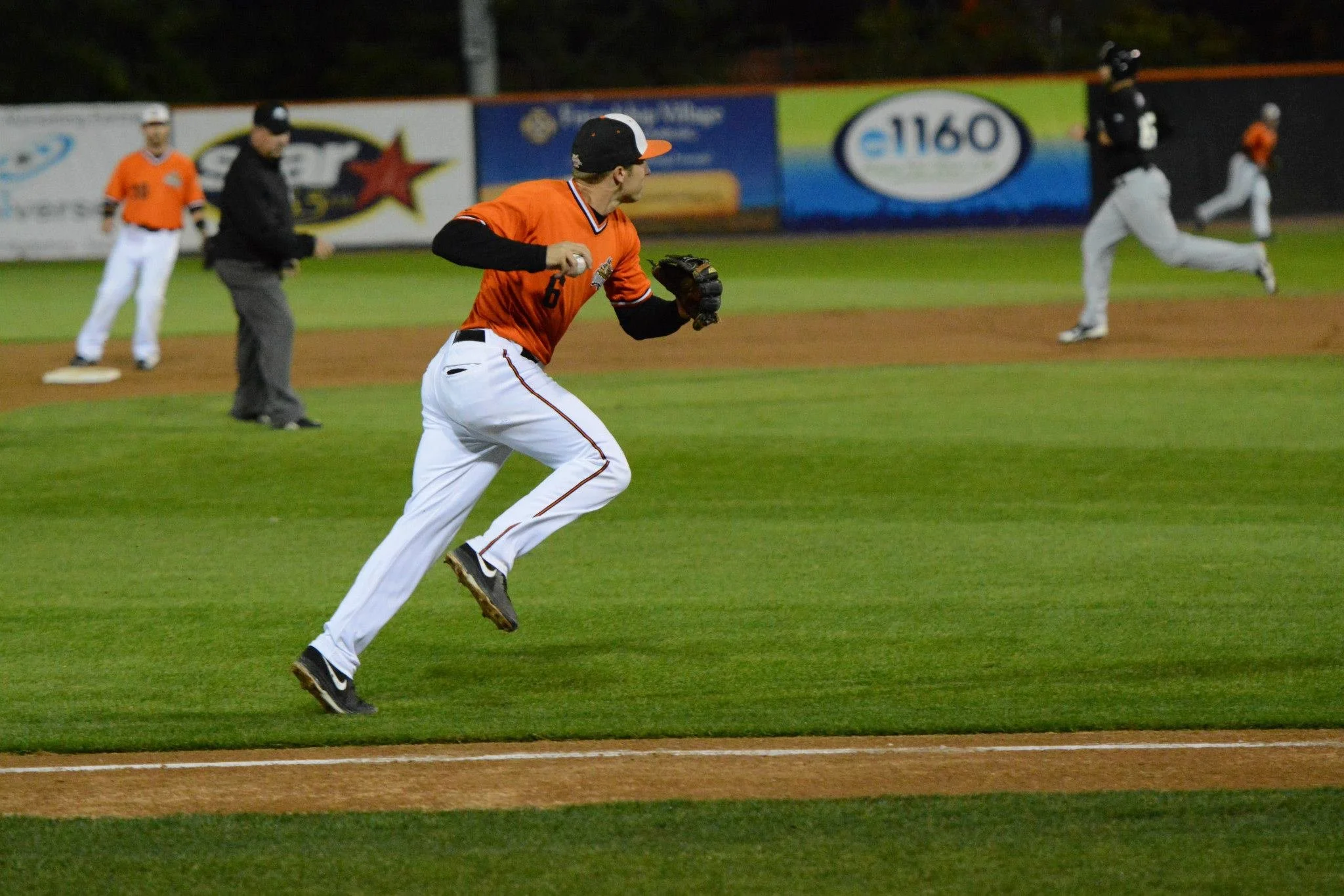A baseball player in orange and white uniform running on the field during a game, with other players and an umpire in the background.