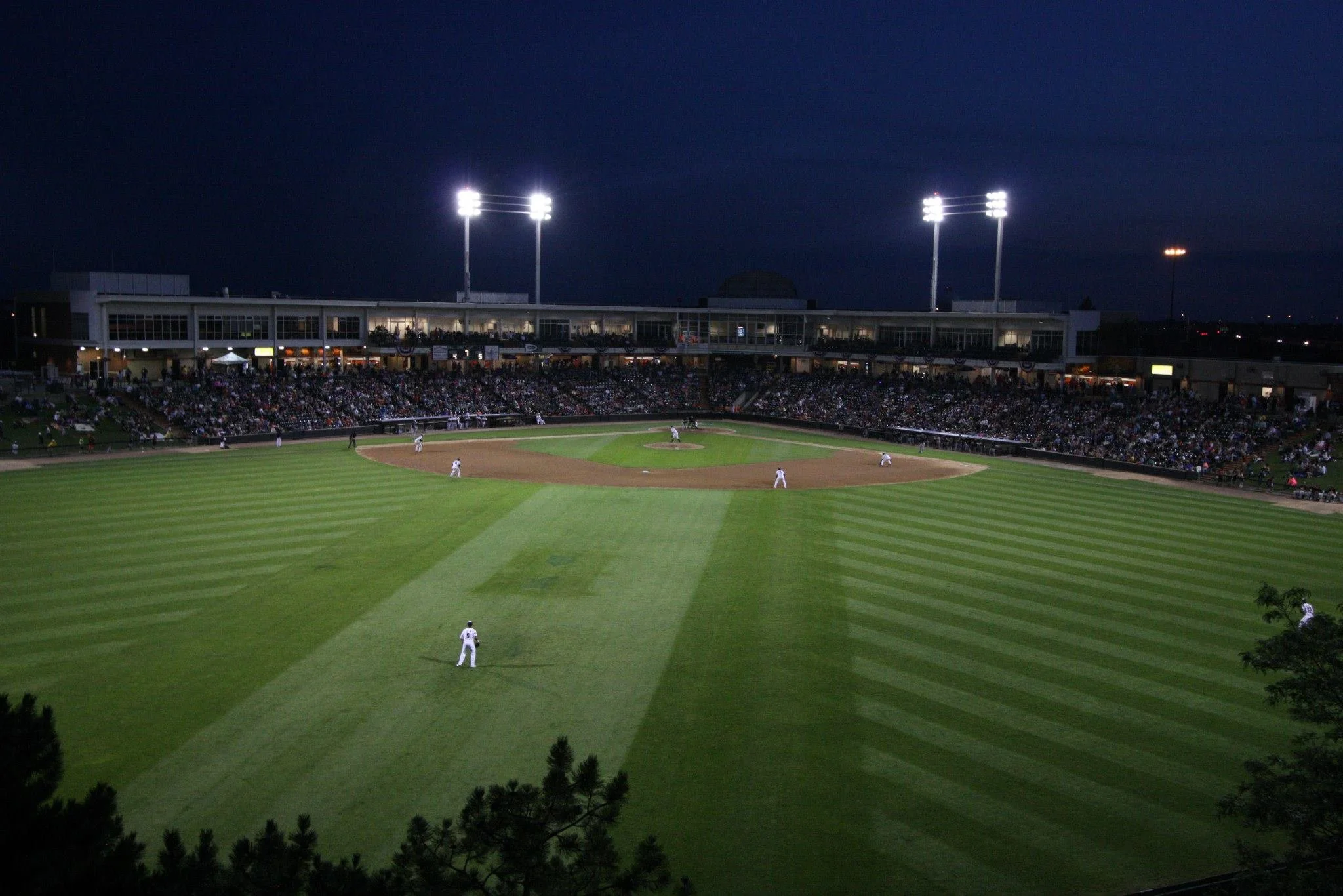Nighttime baseball stadium scene with bright stadium lights illuminating the field and stands filled with fans.