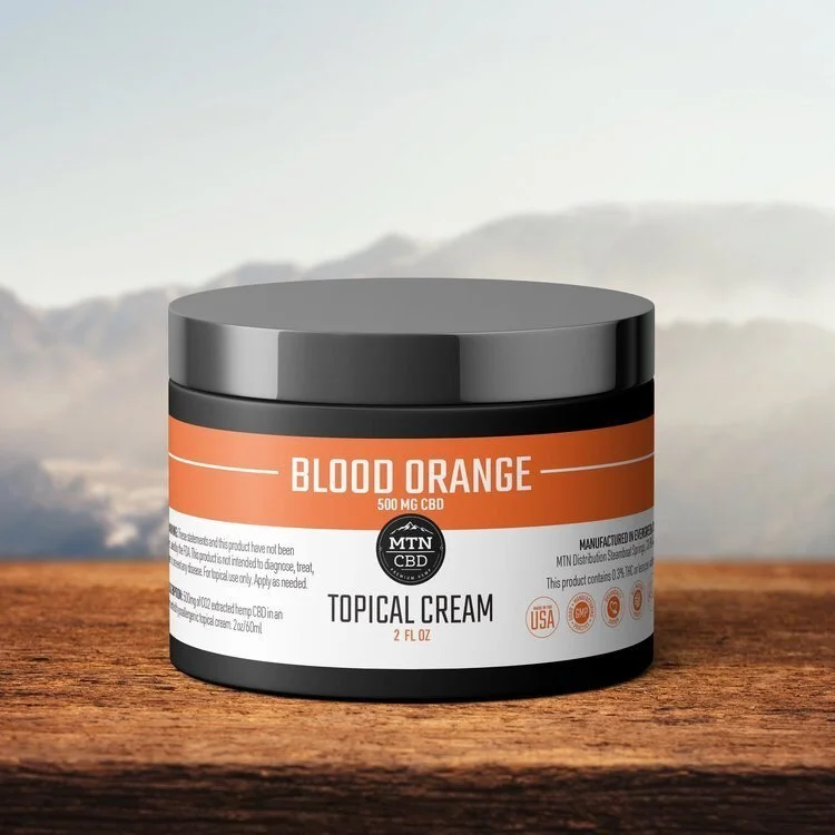 A black jar of blood orange CBD topical cream with an orange and white label, placed on a wooden surface with a blurred outdoor background.
