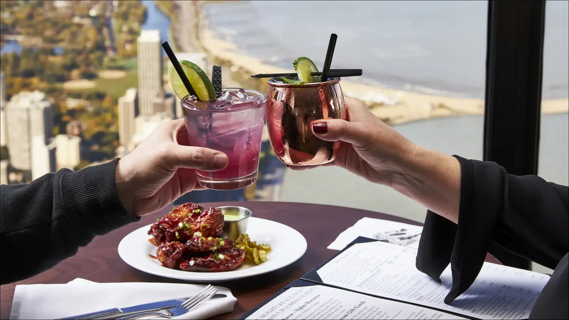 Two people toasting lime-garnished cocktails above a city and waterfront.
