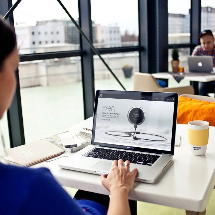 Person sitting at a white desk using a MacBook Pro laptop, displaying a website for Xen wellness products. The desk has a beige notebook, a yellow and white cup, and some papers. In the background, there are large windows with a cityscape view and an