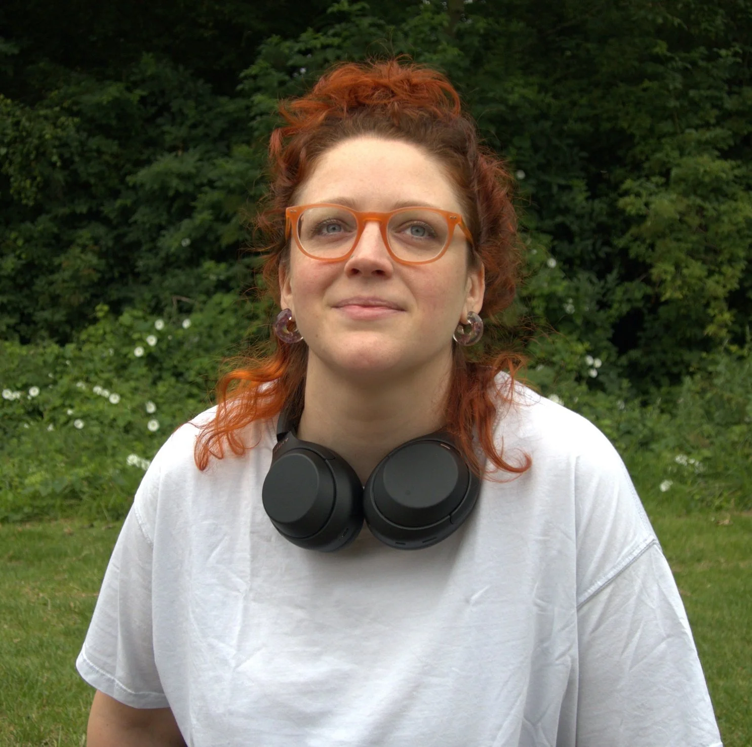 Susannah Worth, founder of Gutsy and host of Loose Threads, sitting in a park with headphones around her neck, wearing orange glasses with henna-orange hair.