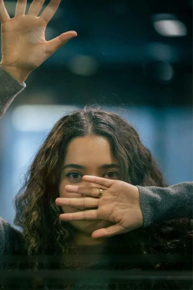 Woman with brown wavy hair covering her face with her flat hand, palm outwards, brown eyes looking directly ahead