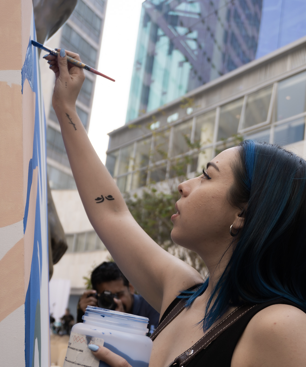 Una mujer con cabello azul pintando en una pared exterior en una ciudad moderna, mientras una persona en el fondo toma una fotografía.